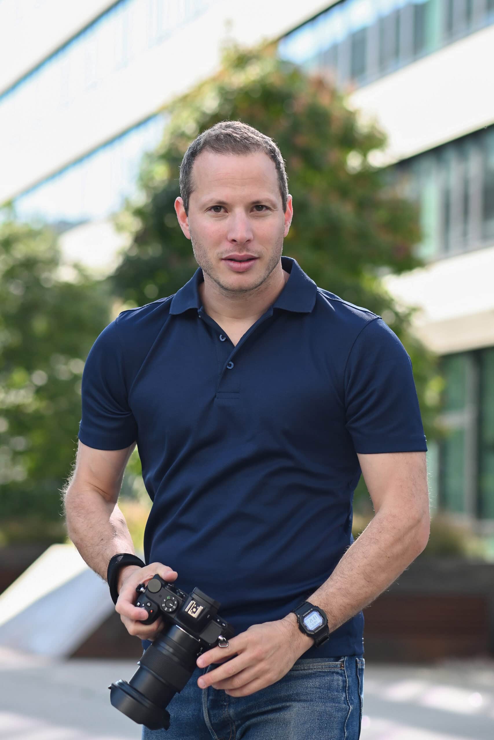 Mann mit Kamera vor moderner Bürogebäudefront, professionelles Portrait, urbane Umgebung, mit Fokus auf Fotografie, Business, Lifestyle, outdoors, leicht sportlich, casual style, natürliche Beleuchtung, Stadtfotografie, kreative Fotokunst.