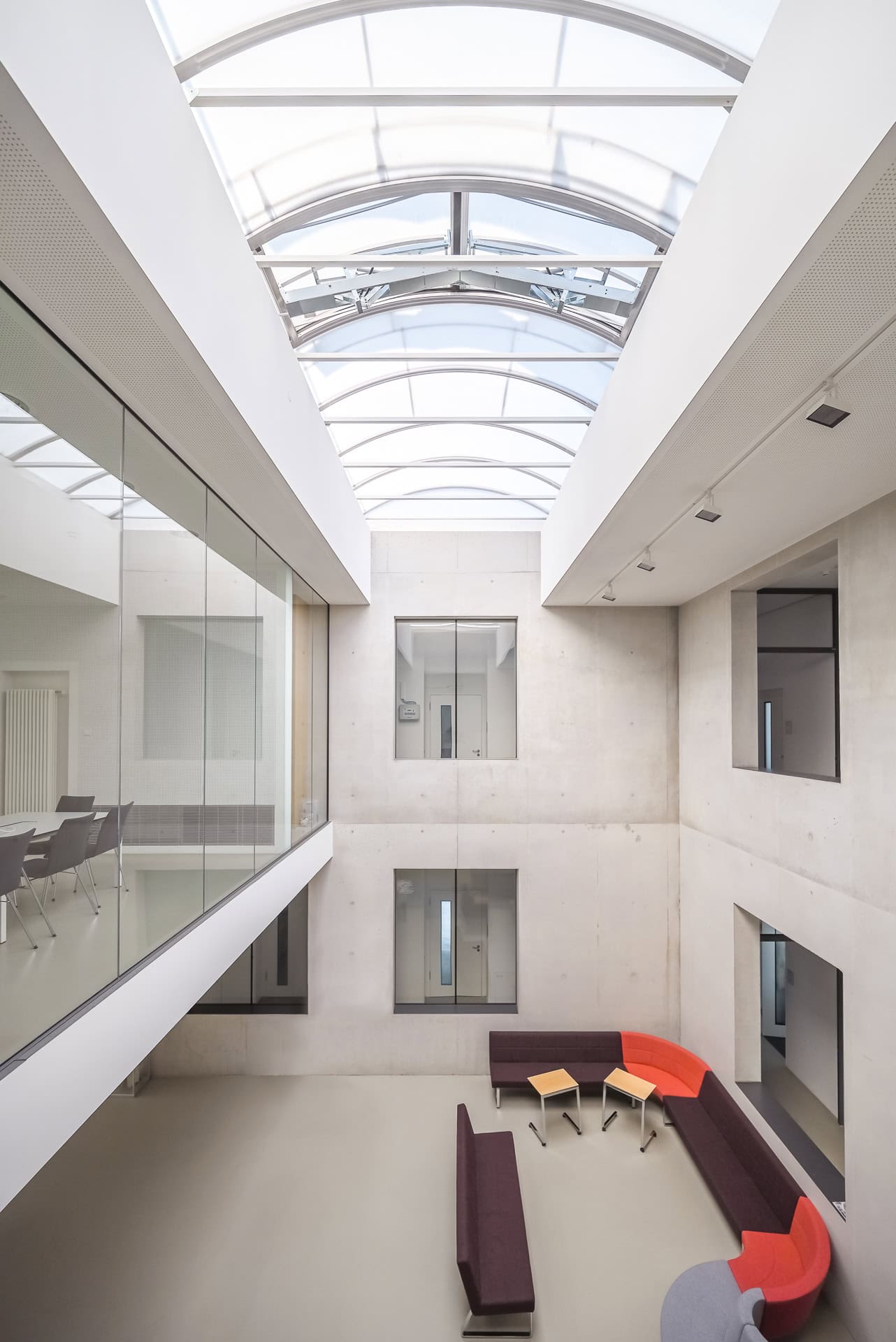 Atrium im Gebäude FIT Materialforschung Freiburg mit Oberlicht, Sichtbetonwänden und farbigen Sitzmöbeln. Architektur von ArGe Architekten, Leins | Ohnemus, Freie Architekten | Part mbB. Foto: Bertrand Weiss, weissfotografie.com.