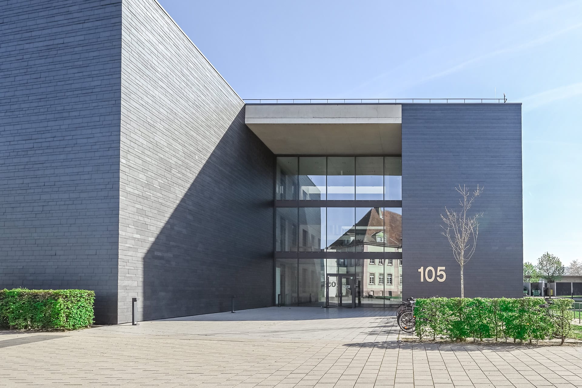 Eingangsbereich von FIT Materialforschung Freiburg mit dunkler Fassade, Glasportal und Hausnummer 105. Architektur von ArGe Architekten, Leins | Ohnemus, Freie Architekten | Part mbB. Foto: Bertrand Weiss, weissfotografie.com.