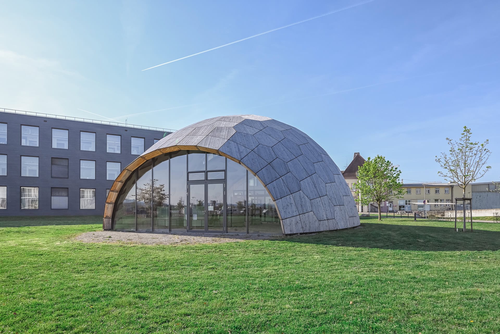 Kuppelförmiger Holzbau von FIT Materialforschung Freiburg mit Forschungsgebäude im Hintergrund. Architektur von ArGe Architekten, Leins | Ohnemus, Freie Architekten | Part mbB. Foto: Bertrand Weiss, weissfotografie.com.