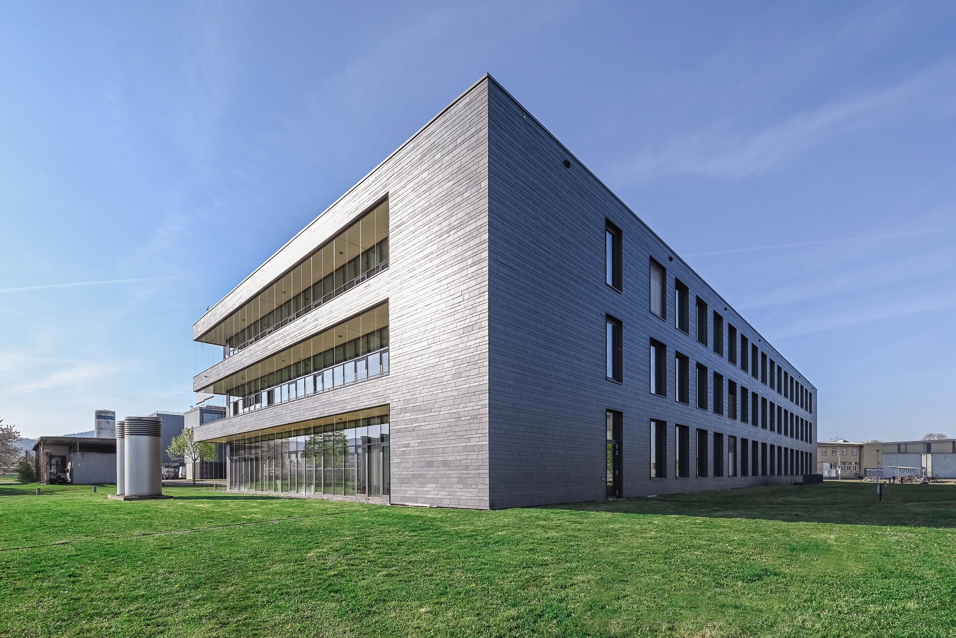 Außenansicht von FIT Materialforschung Freiburg mit grauem Baukörper, Glasfassade und Grünfläche. Architektur von ArGe Architekten, Leins | Ohnemus, Freie Architekten | Part mbB. Foto: Bertrand Weiss, weissfotografie.com.