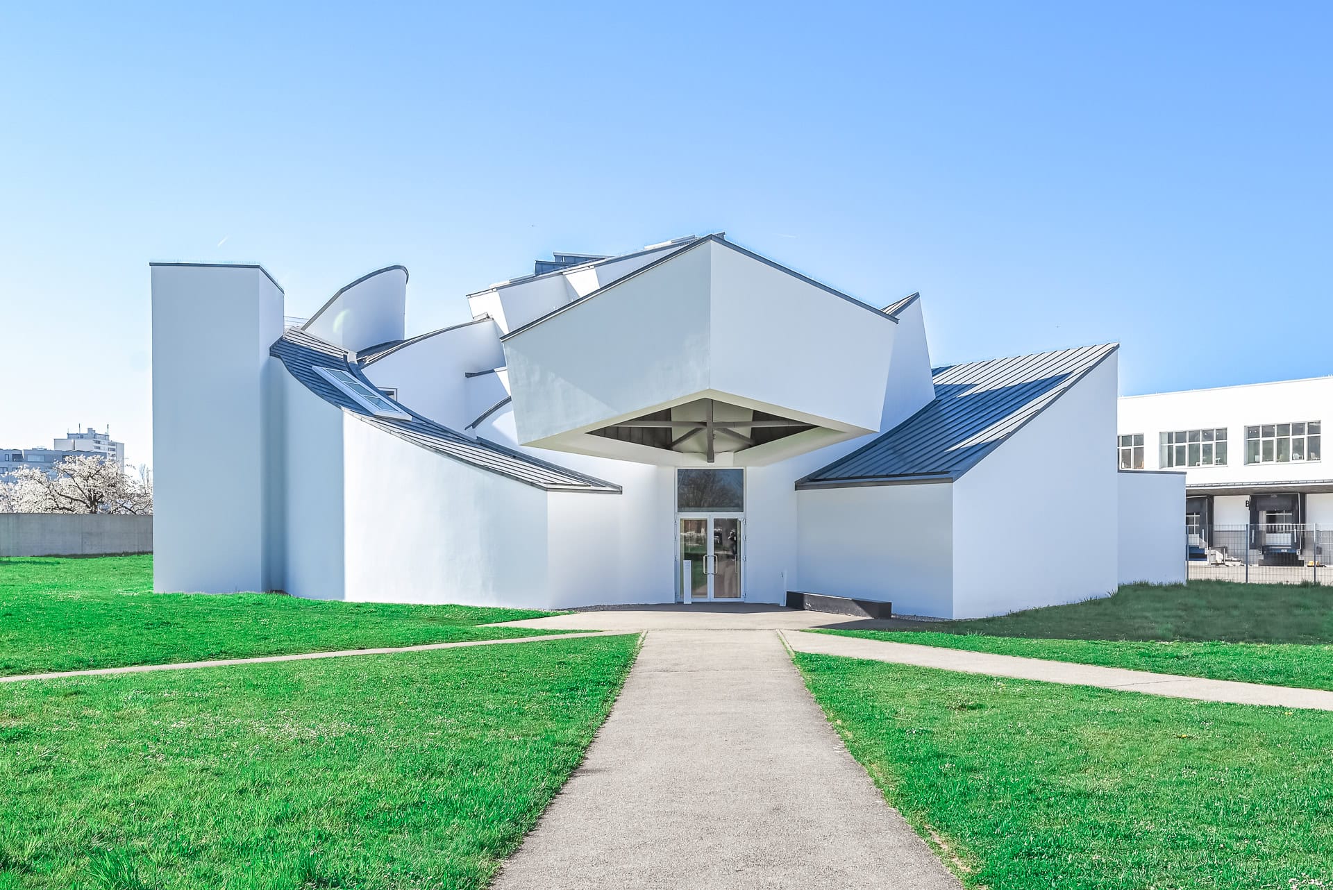 Frontansicht des Vitra Design Museums mit weißer Fassade, gefalteten Baukörpern und Weg durch die Grünfläche. Architektur von Frank O. Gehry. Foto: Bertrand Weiss, weissfotografie.com.