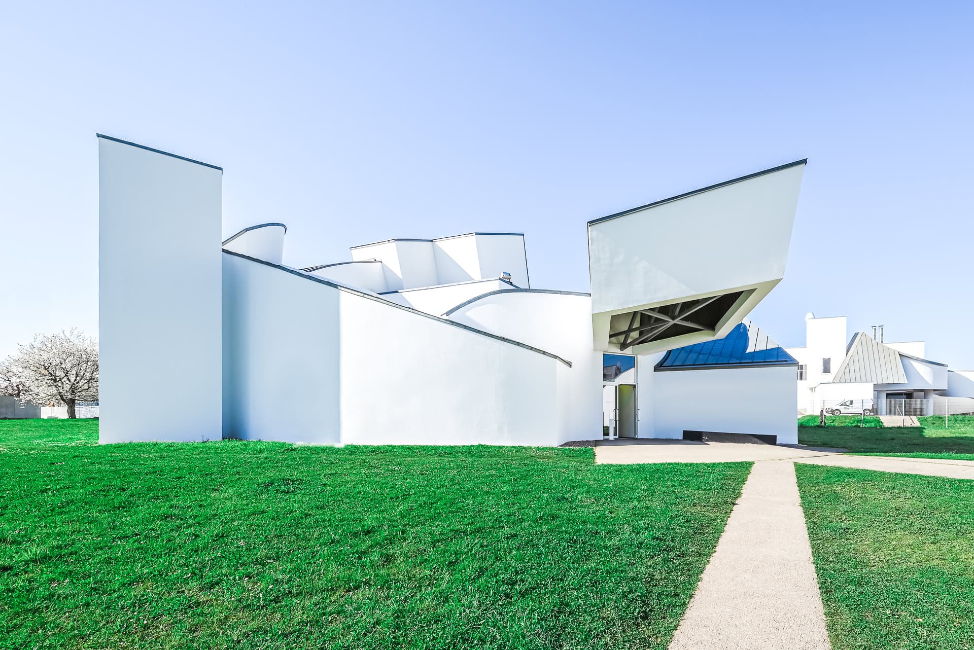 Seitliche Außenansicht des Vitra Design Museums mit weißen Volumen, gebogenen Dachlinien und Eingang. Architektur von Frank O. Gehry. Foto: Bertrand Weiss, weissfotografie.com.