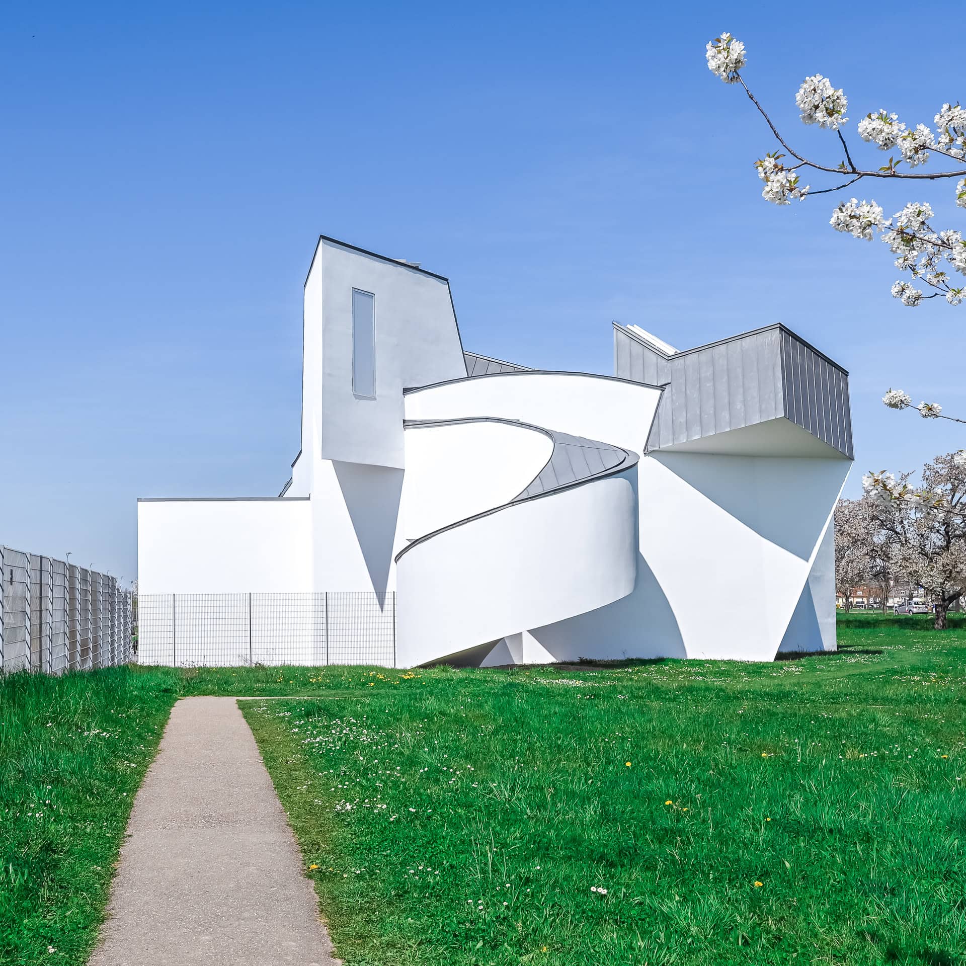 Außenansicht des Vitra Design Museums mit geschwungenem Baukörper, weißen Fassaden und blühenden Bäumen. Architektur von Frank O. Gehry. Foto: Bertrand Weiss, weissfotografie.com.