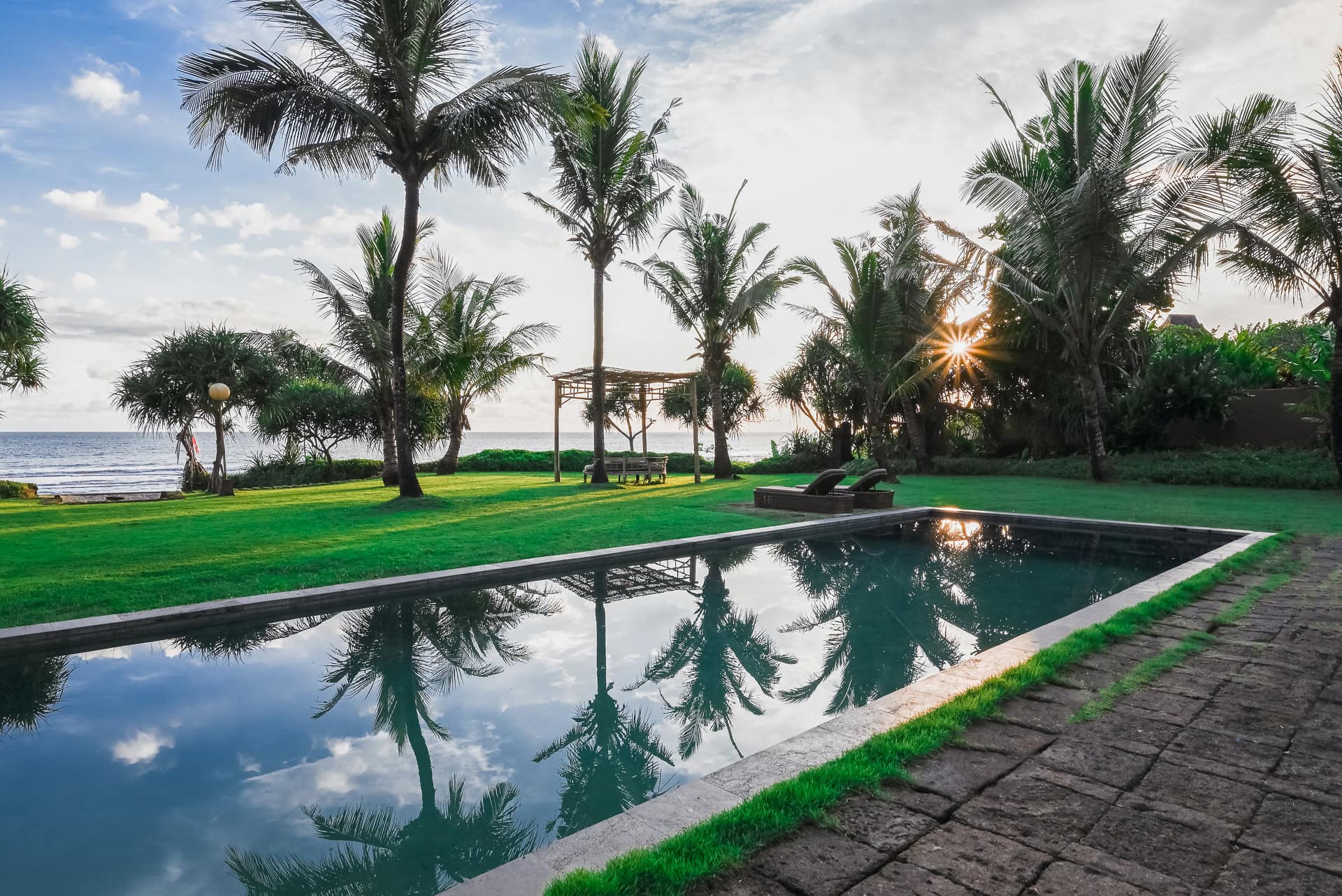 Poolanlage des WakaGangga Resort zwischen Palmen mit tief stehender Sonne. Architektur von I Ketut Siandana. Foto: Bertrand Weiss, weissfotografie.com.