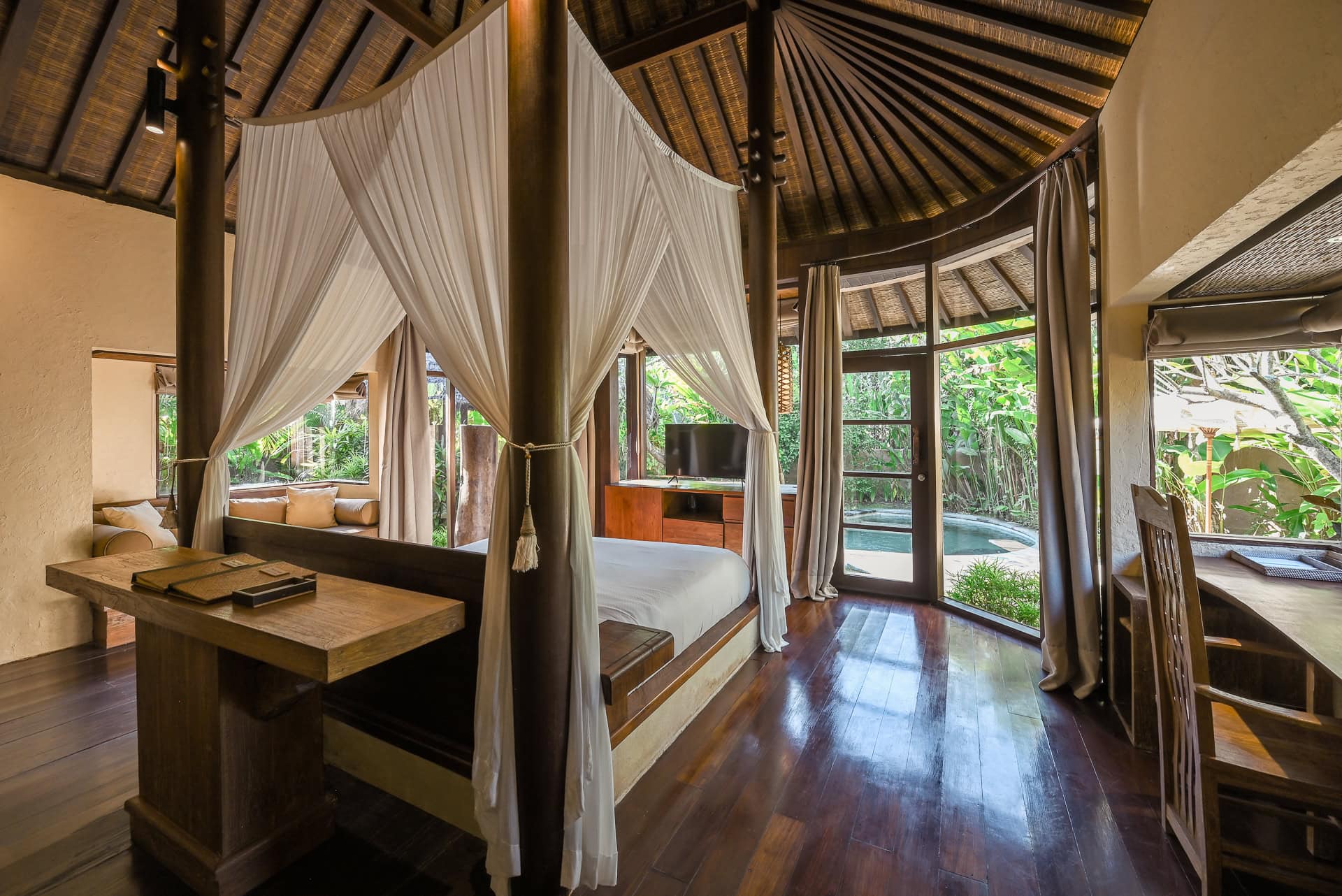 Schlafraum einer Villa mit Himmelbett, dunklem Holz, Reetdach und Blick zum privaten Pool. Architektur von I Ketut Siandana. Foto: Bertrand Weiss, weissfotografie.com.