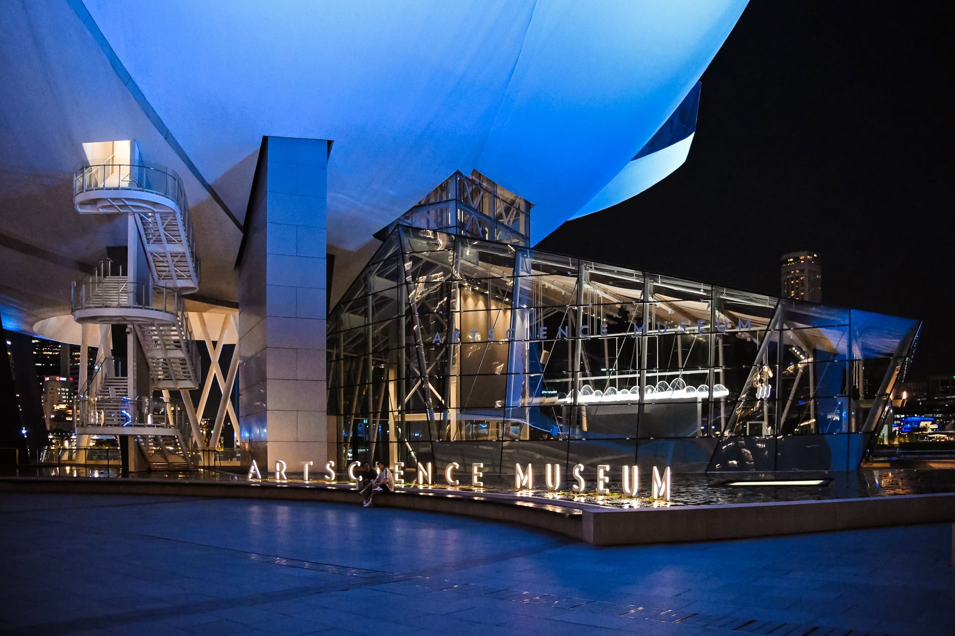 Beleuchteter Eingangsbereich des ArtScience Museum bei Nacht mit Glasfassade und auskragenden weißen Baukörpern. Architektur von Safdie Architects. Foto: Bertrand Weiss, weissfotografie.com.
