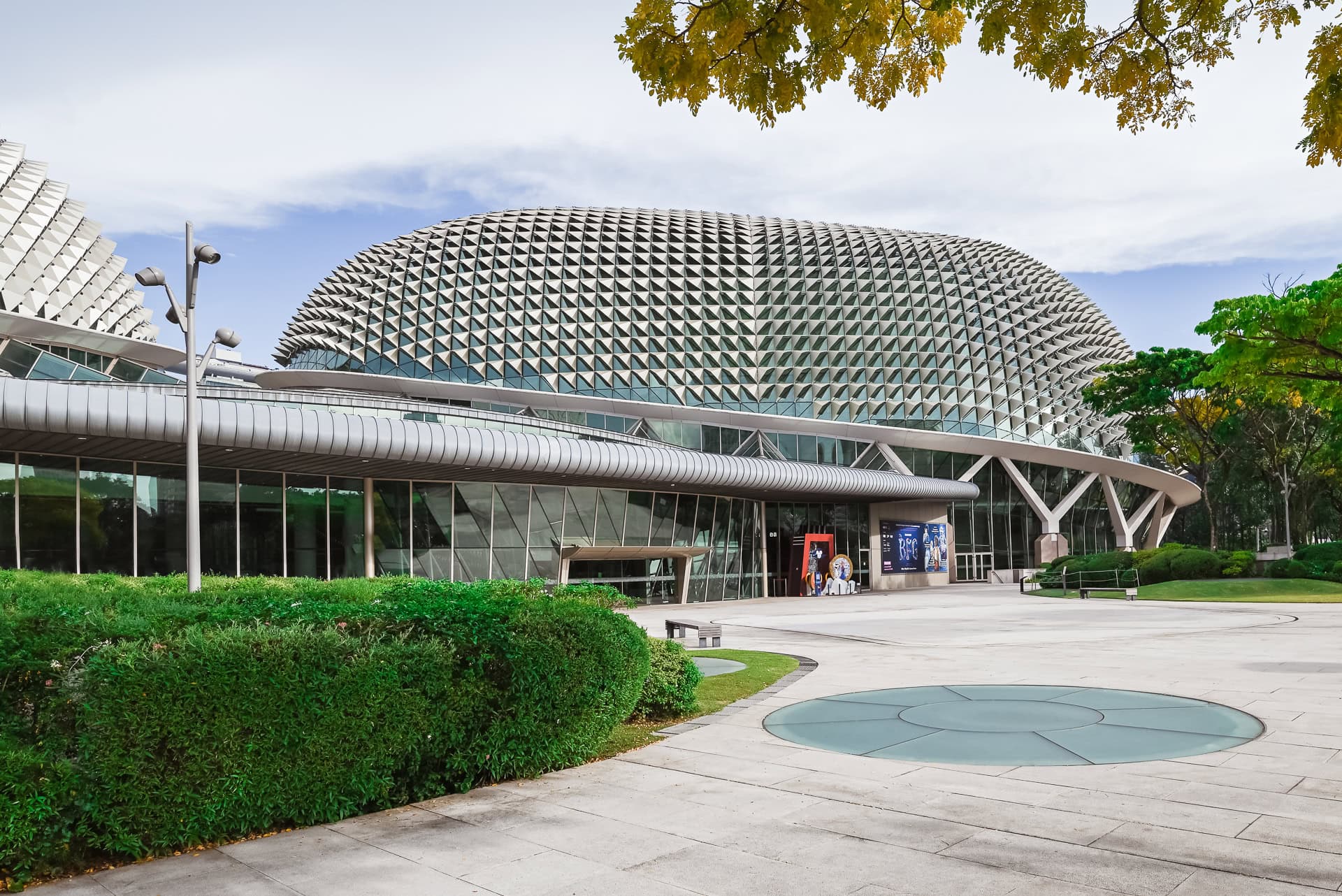 Weitläufiger Vorplatz des Esplanade mit geschwungener Fassade, Begrünung und kreisförmigem Glaselement im Boden. Architektur von DP Architects und Michael Wilford & Partners. Foto: Bertrand Weiss, weissfotografie.com.