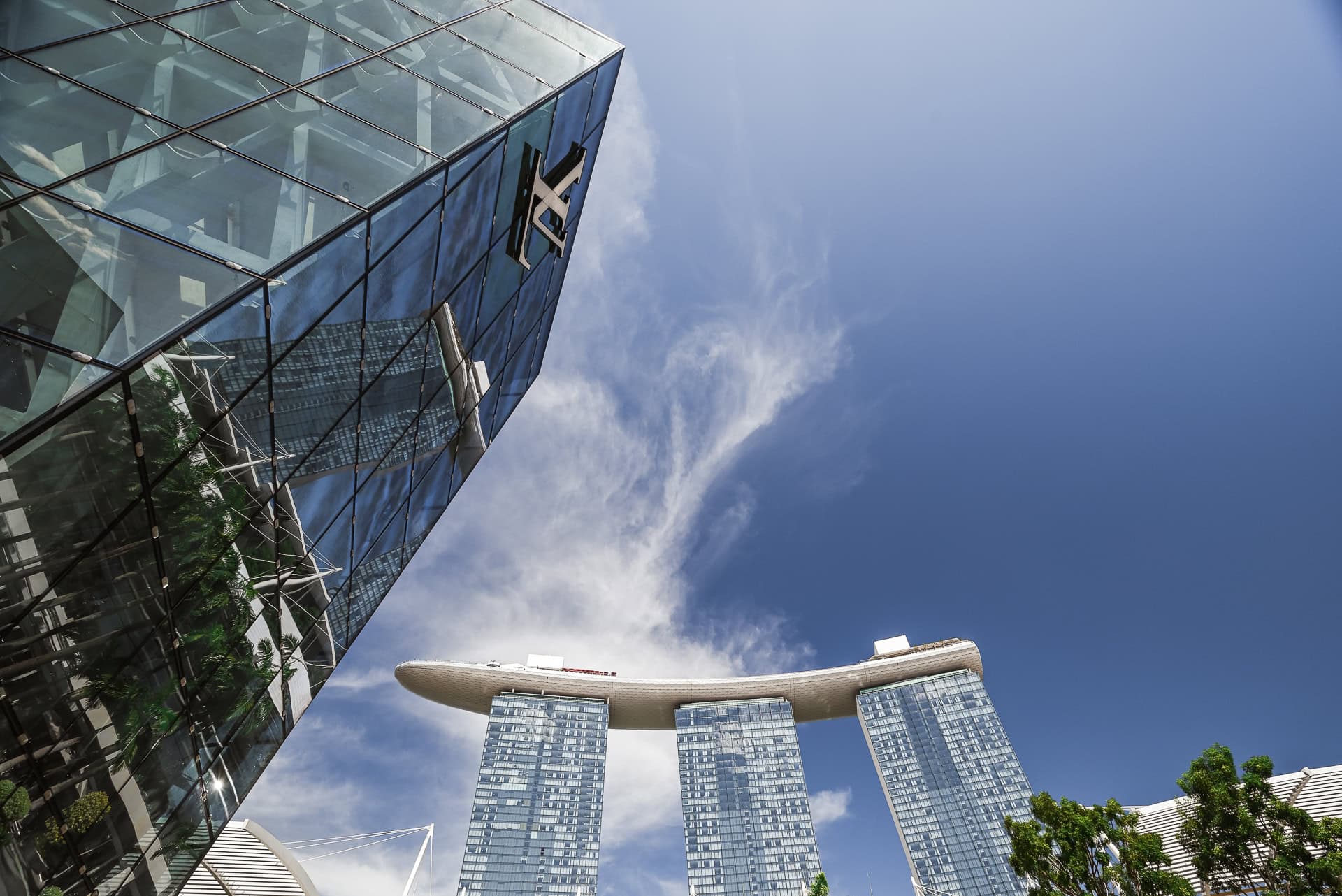 Gläserner Louis Vuitton Island Maison Pavillon mit Marina Bay Sands im Hintergrund unter blauem Himmel. Architektur von Safdie Architects. Foto: Bertrand Weiss, weissfotografie.com.
