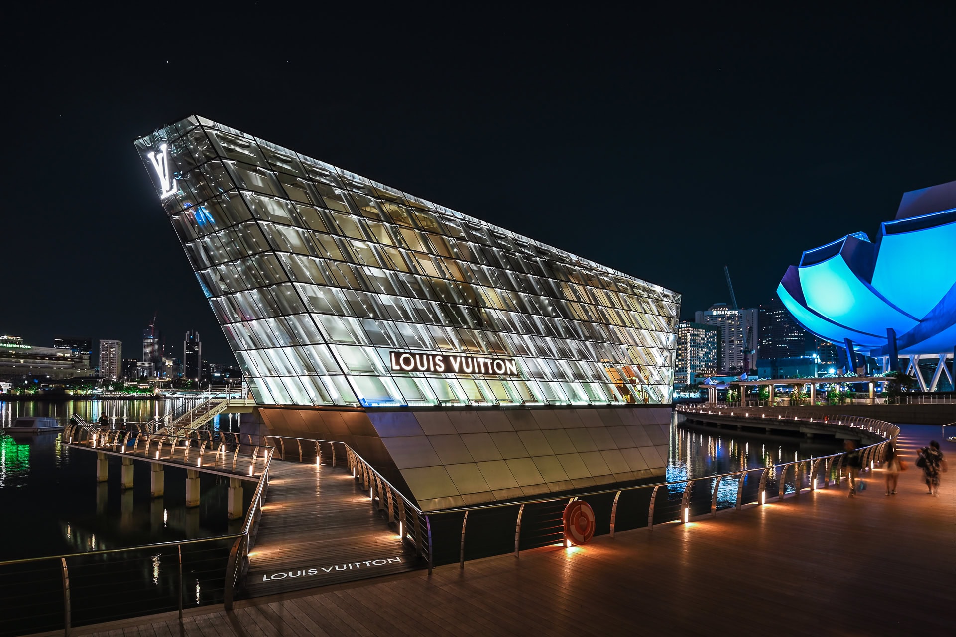 Nächtliche Seitenansicht des Louis Vuitton Island Maison mit beleuchteter Glasfassade und Stegen über dem Wasser. Architektur von Safdie Architects. Foto: Bertrand Weiss, weissfotografie.com.