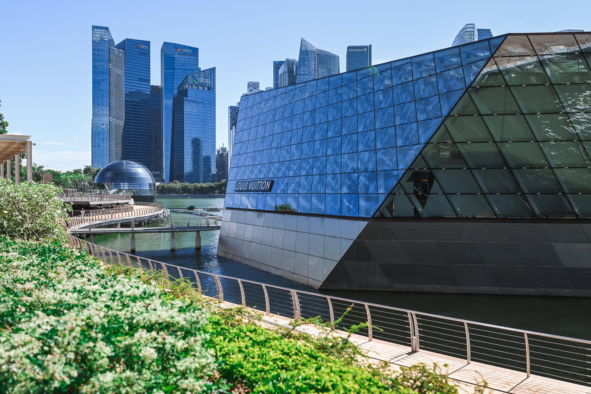 Seitliche Ansicht des Louis Vuitton Pavillons mit spiegelnder Glasfassade, Uferpromenade und Hochhäusern der Marina Bay. Architektur von Safdie Architects. Foto: Bertrand Weiss, weissfotografie.com.